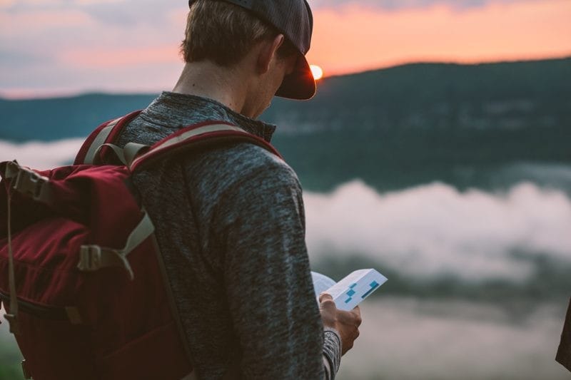 chico rubio de espaldas con gorra y con una mochila de color roja mirando un libro. Enfrente de él, la puesta de sol